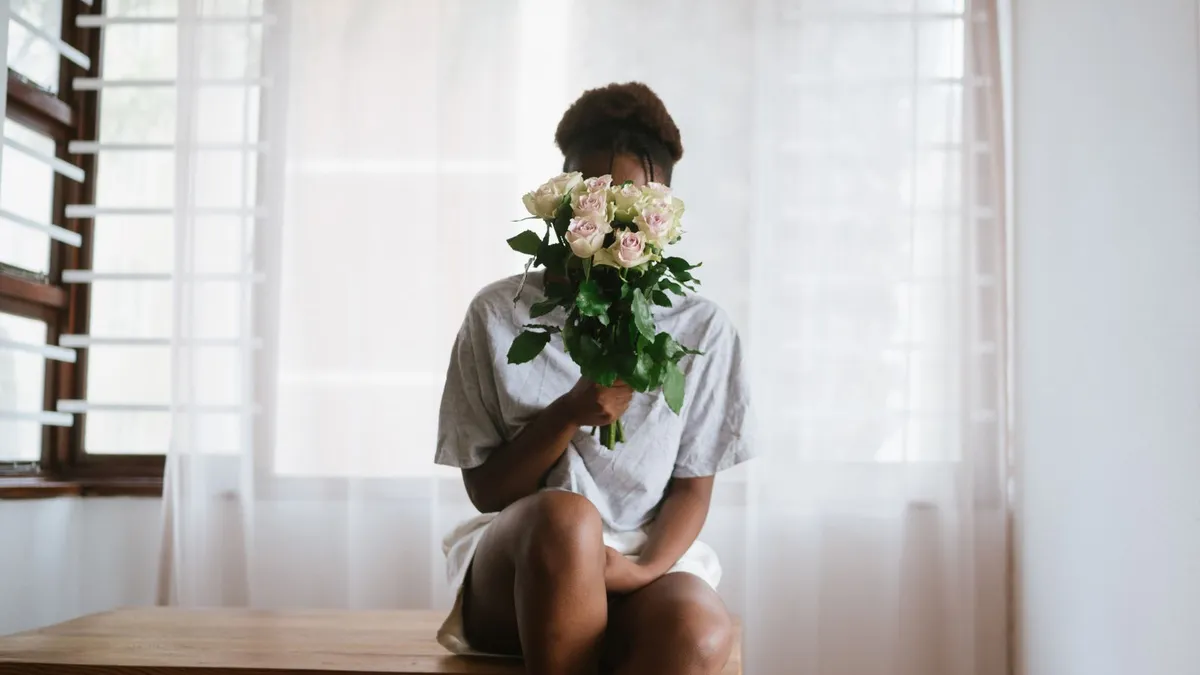 Eine Person sitzt am Fenster auf einer Holzbank und hält einen Strauß heller Rosen vor das Gesicht