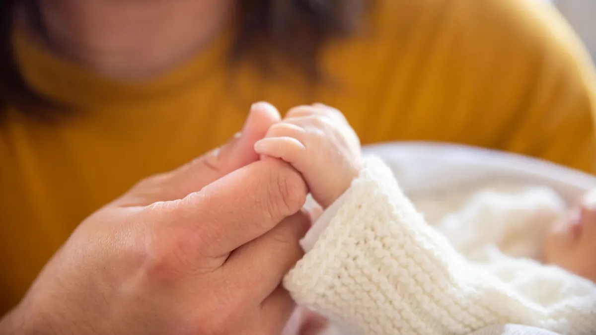 Eine Erwachsenenhand hält die kleine Hand eines Neugeborenen im weißen Strickjäckchen