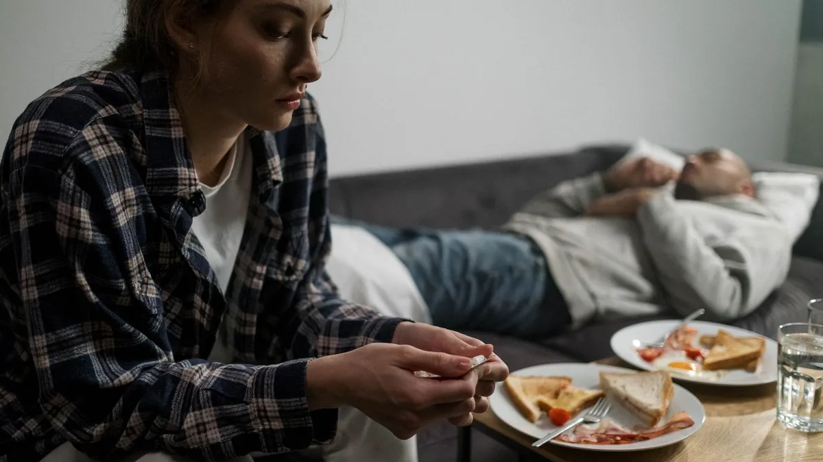 Eine Frau sitzt nachdenklich vor einem unberührten Frühstück, hinter ihr liegt ein Mann abgewandt auf dem Sofa