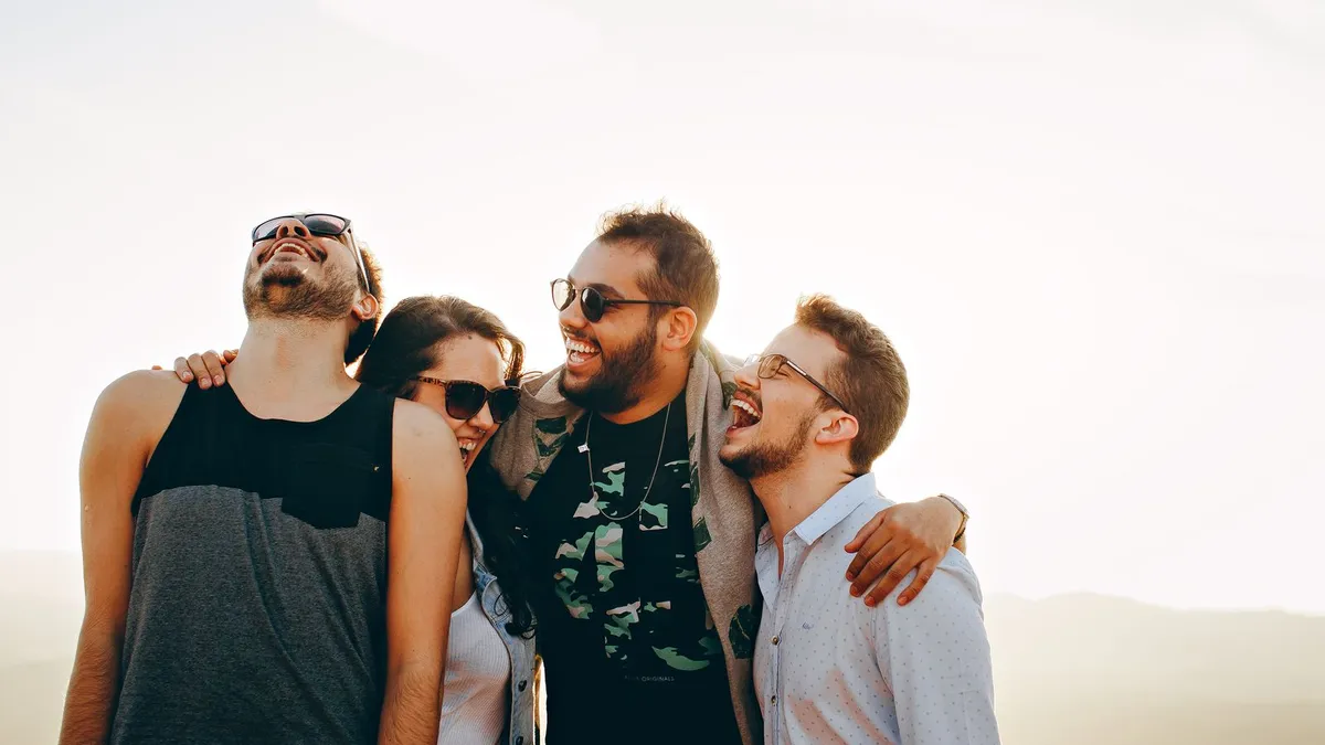 Vier Freunde lachen im Gegenlicht und halten sich im Arm, mehrere tragen Sonnenbrillen