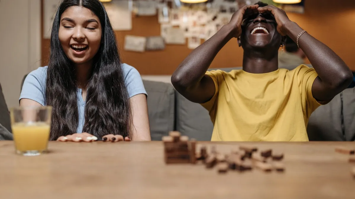 Zwei Freunde lachen am Holztisch über einen eingestürzten Turm aus Holzklötzen