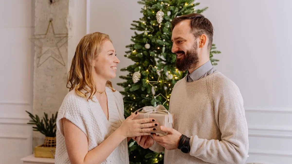 Frau und Mann mit Bart übergeben sich vor einem beleuchteten Weihnachtsbaum lächelnd ein kleines Geschenk