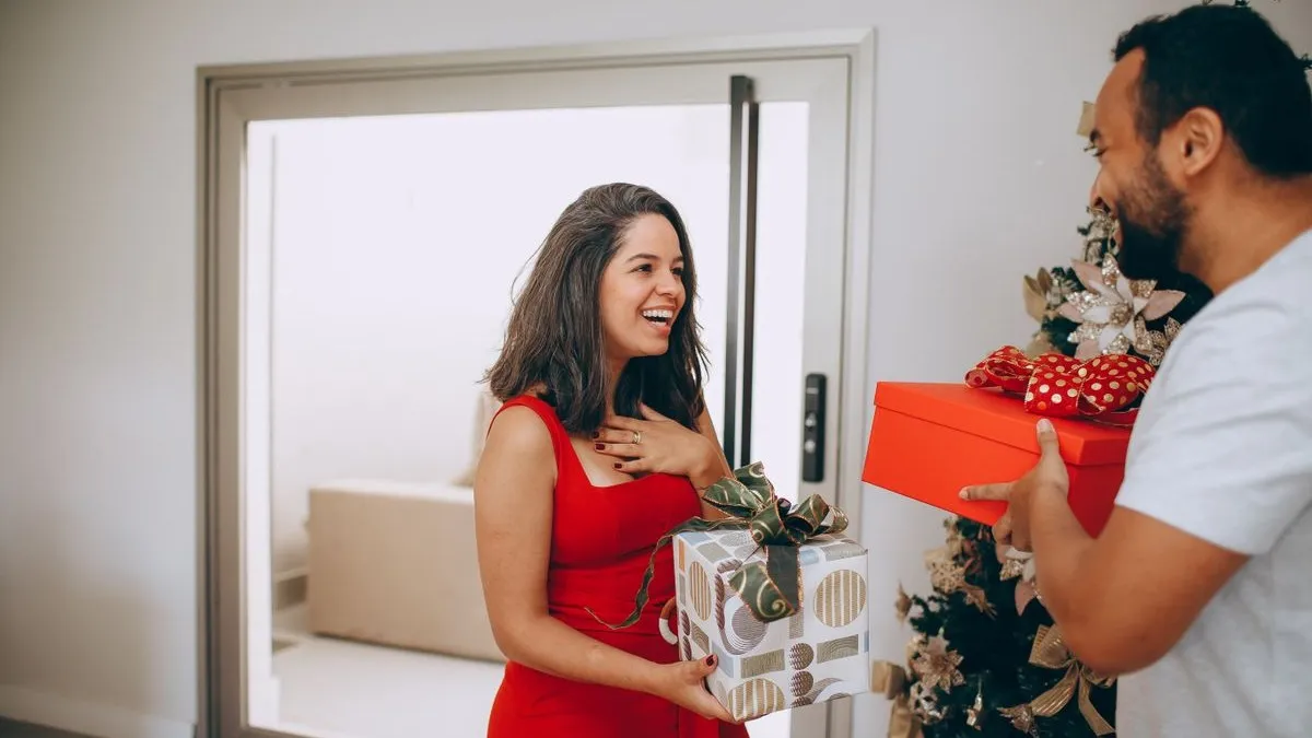 Lachende Frau im roten Kleid bekommt vor dem Weihnachtsbaum ein rotes Geschenk von ihrem Partner