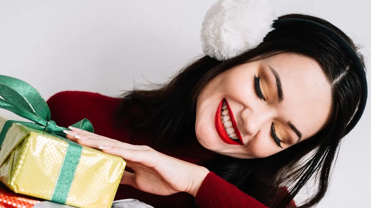 Lachende Frau mit Pompon-Haarreif legt beide Hände auf ein grün verpacktes Weihnachtsgeschenk
