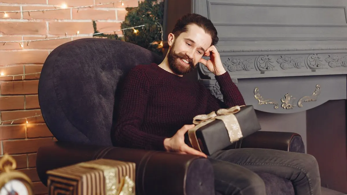 Lächelnder Mann mit Bart sitzt in einem Sessel am Kamin und hält ein verpacktes Geschenk