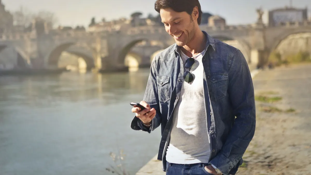 Lächelnder Mann im Jeanshemd schaut auf sein Smartphone, im Hintergrund ein Fluss mit einer Steinbrücke