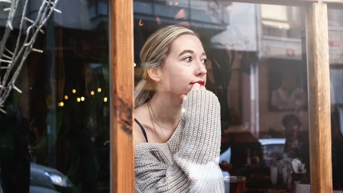 Junge Frau blickt nachdenklich durch ein Café-Fenster, die Hand am Kinn